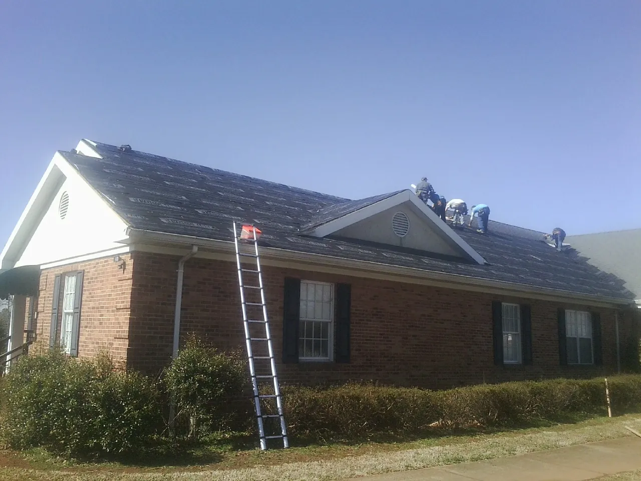 Crew tearing shingles off a brick ranch roof with a ladder set — Emergency Roof Repair work by our crew in Comstock