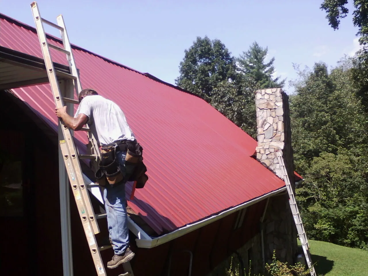 Roofer on a ladder beside a red metal roof and stone chimney — Soffit & Fascia Repair by Roofing Brothers in Comstock